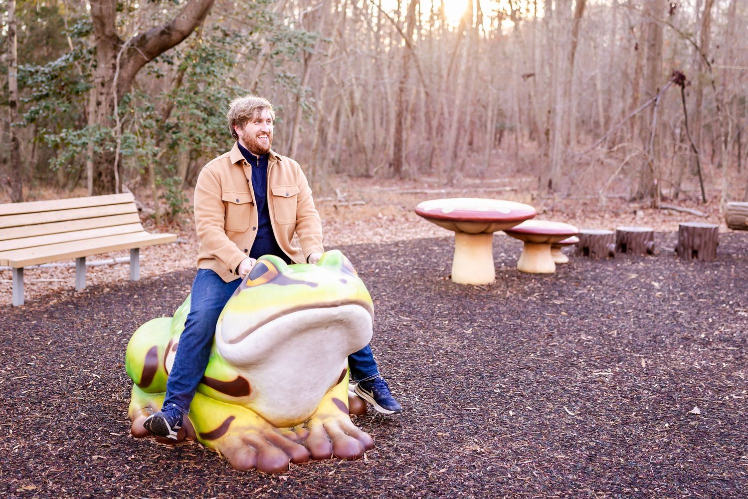 A figure in a tan jacket and blue pants sits on a large decorative frog statue in an outdoor park setting at sunset.