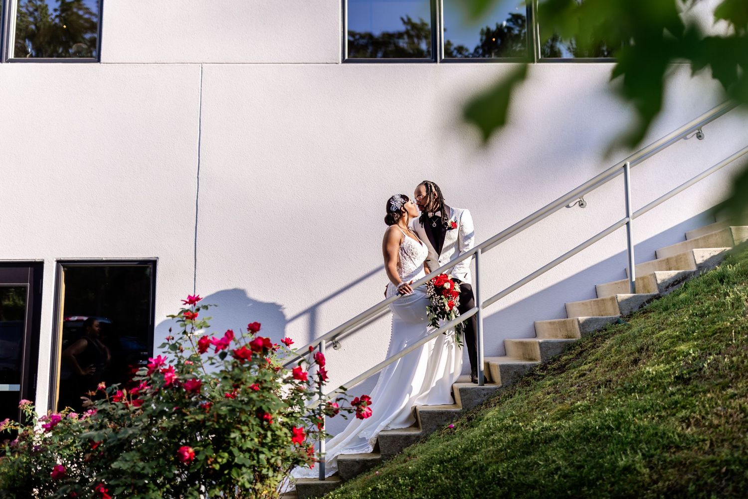 Bride and groom standing on modern staircase surrounded by blooming red roses.