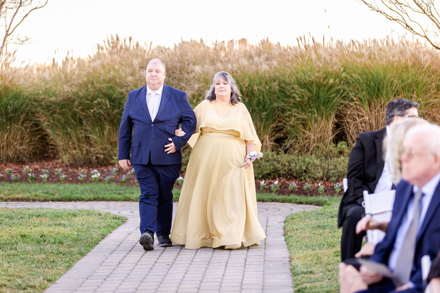Formal procession along garden pathway with attendants wearing blue suit and gold gown.