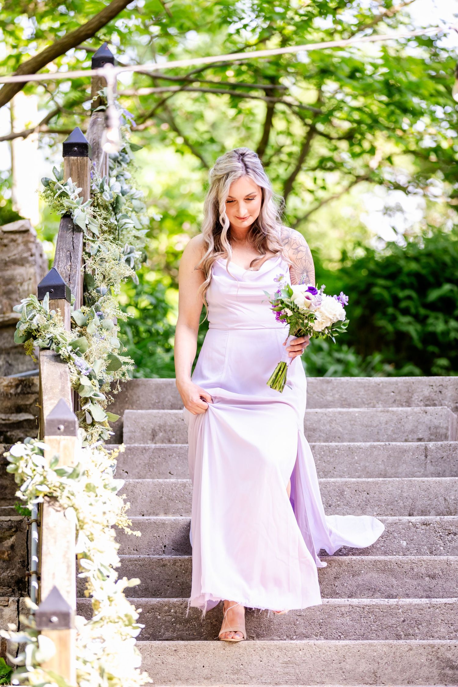 Woman in flowing pink dress poses with bouquet on stone staircase adorned with white flowers.
