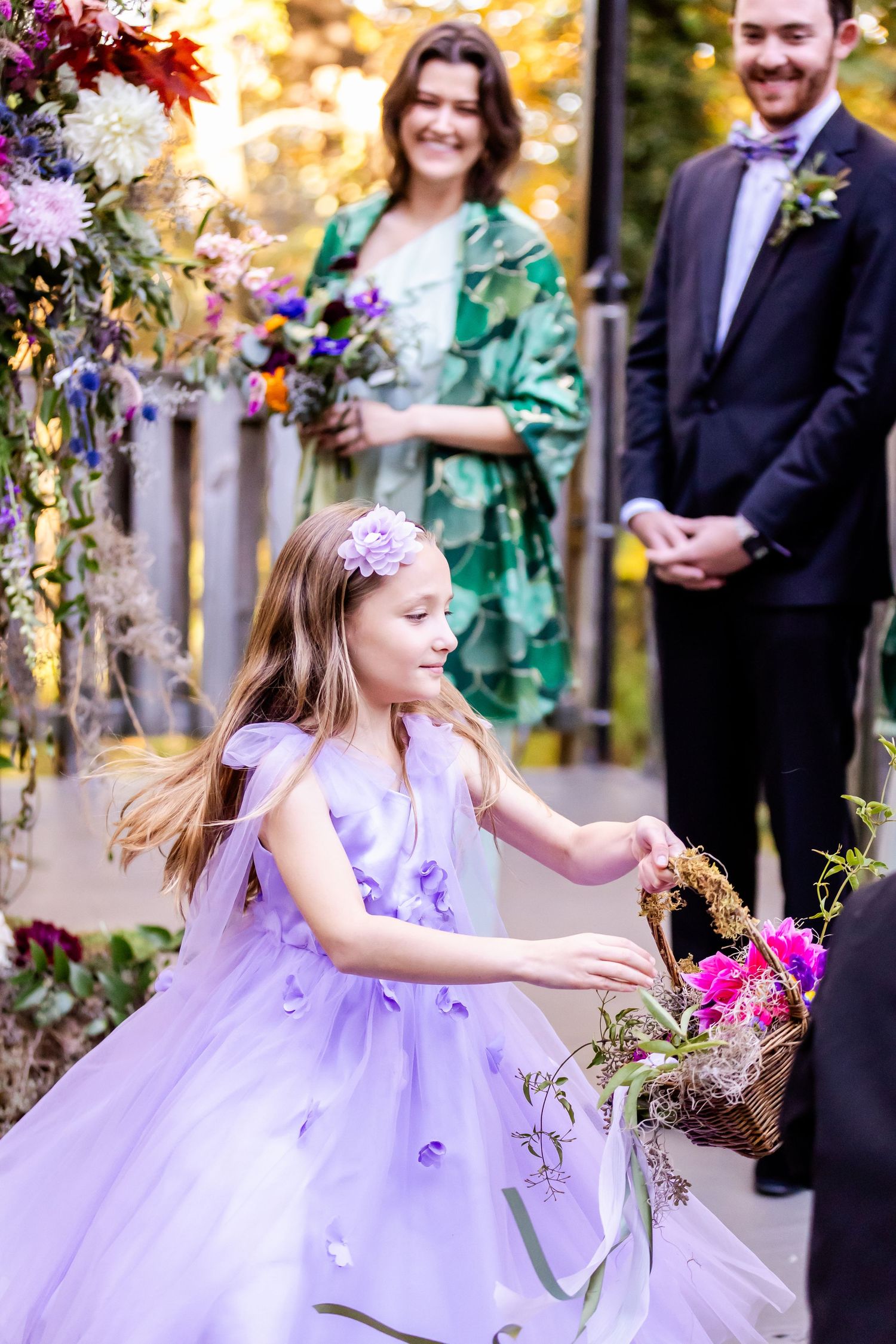 A young flower girl in a purple tulle dress dances under a floral arch during an outdoor wedding ceremony.