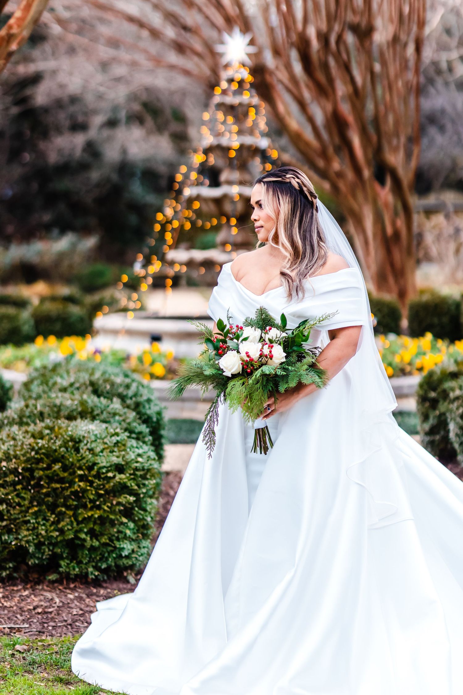 A bride in an off-shoulder white gown holds a floral bouquet in a garden with a lit Christmas tree in the background.