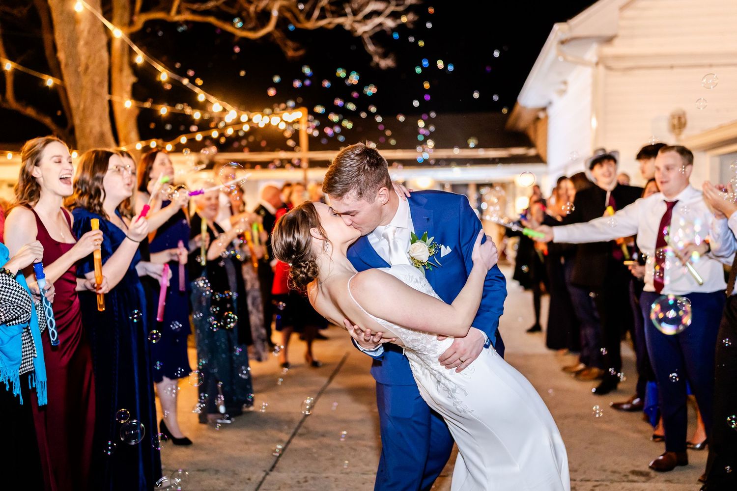 Wedding party dancing and celebrating outdoors under twinkling lights at night.