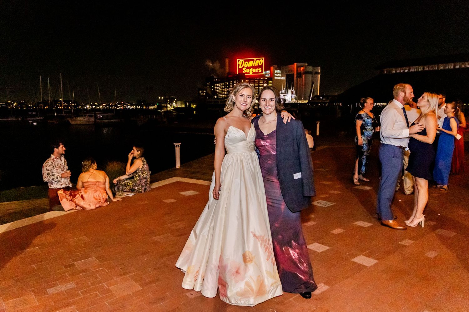 Wedding couple poses during evening reception on waterfront patio with city lights in background.
