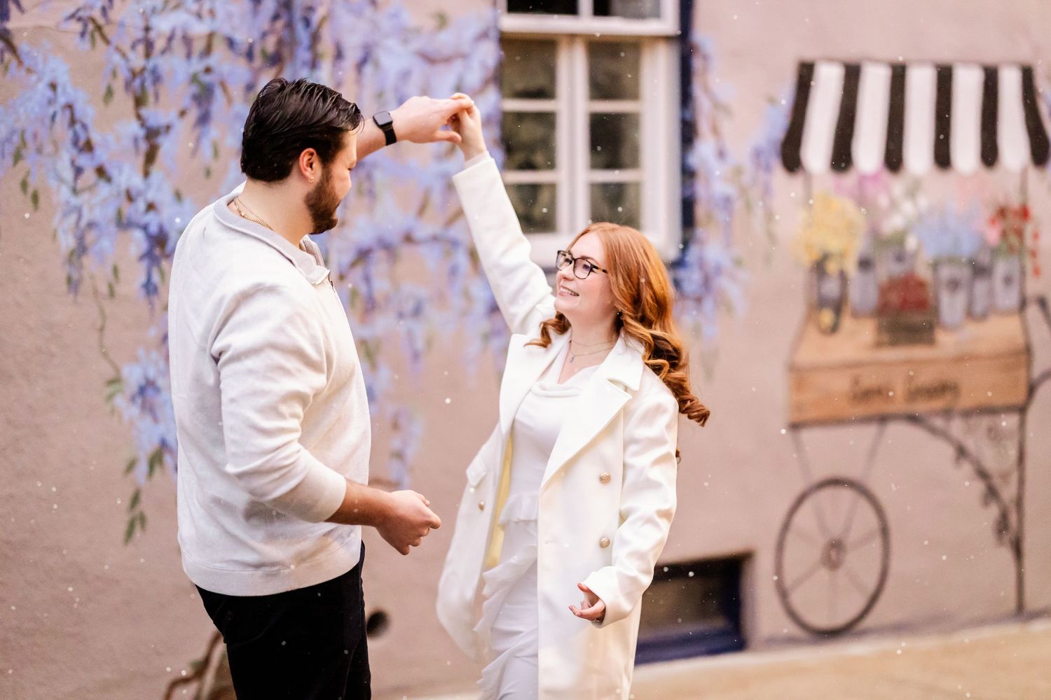 A couple in white outfits dance together outdoors against a pink wall with hanging wisteria flowers.