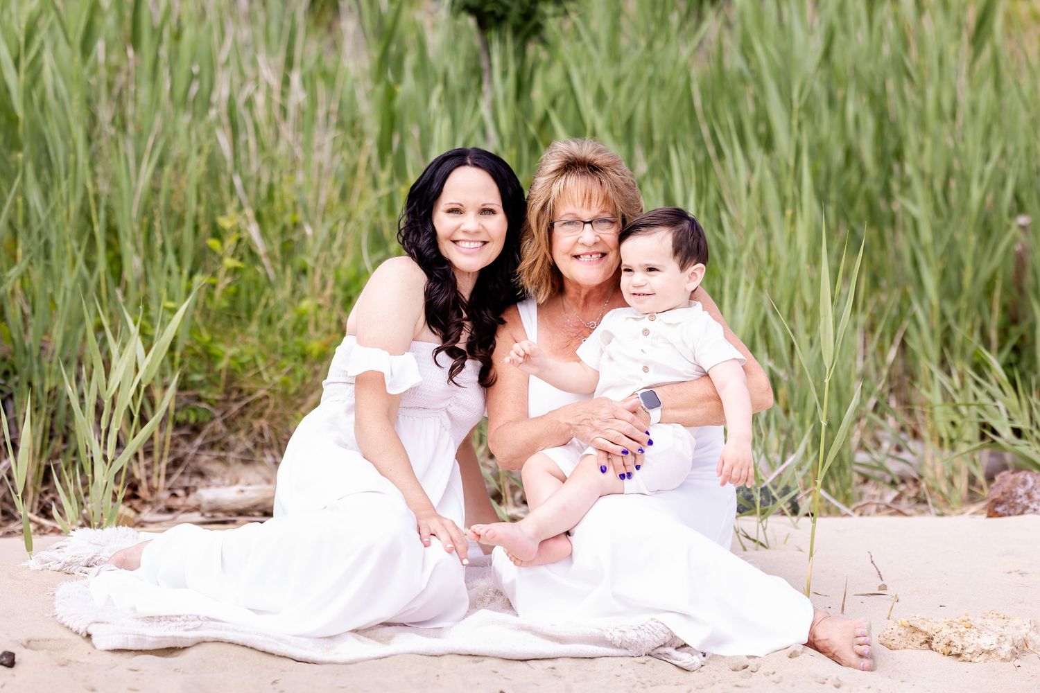 Three people wearing white outfits sit together on a sandy beach with tall grass in the background.
