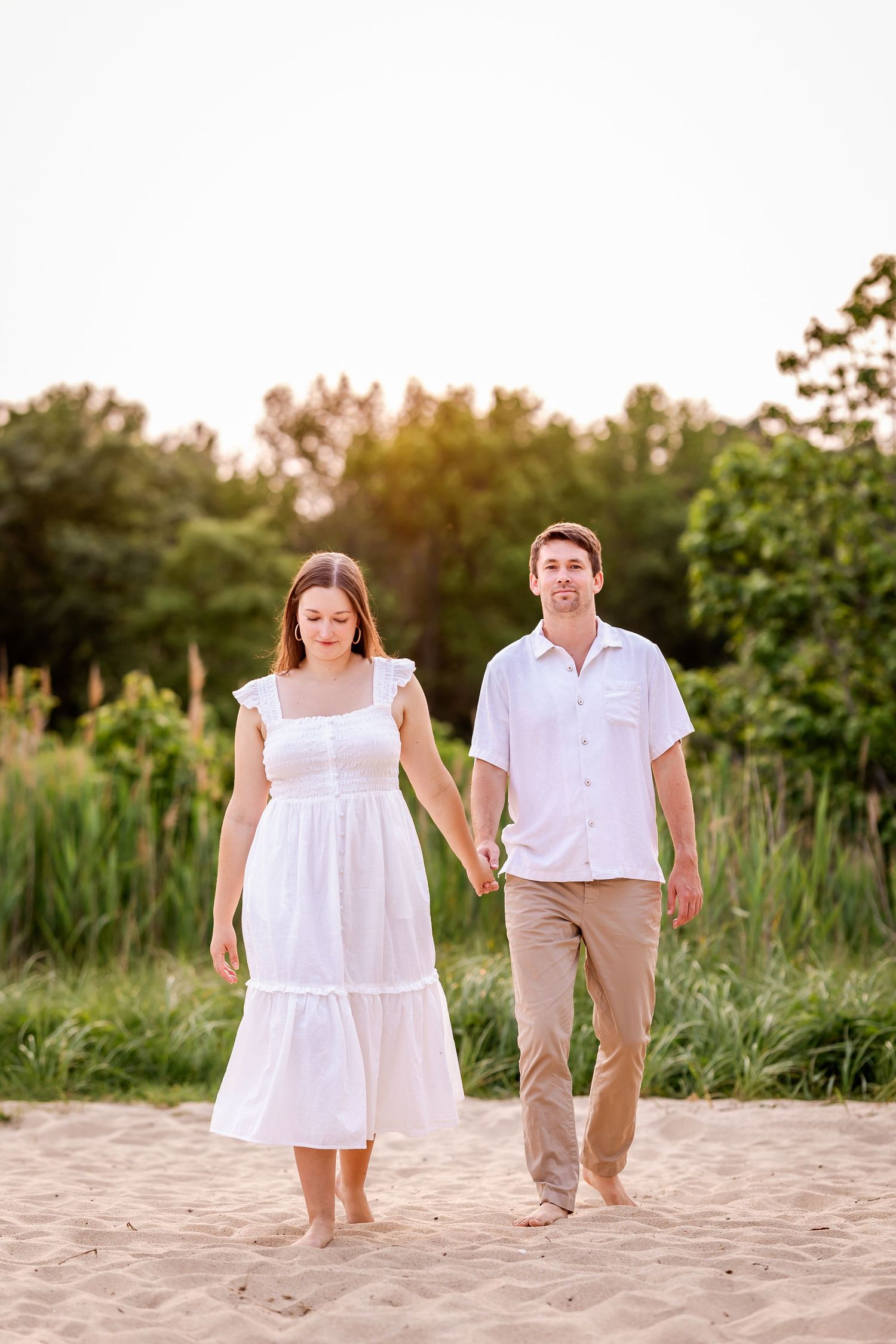 Child and parent walking hand in hand on sandy beach at sunset wearing white and khaki clothing.