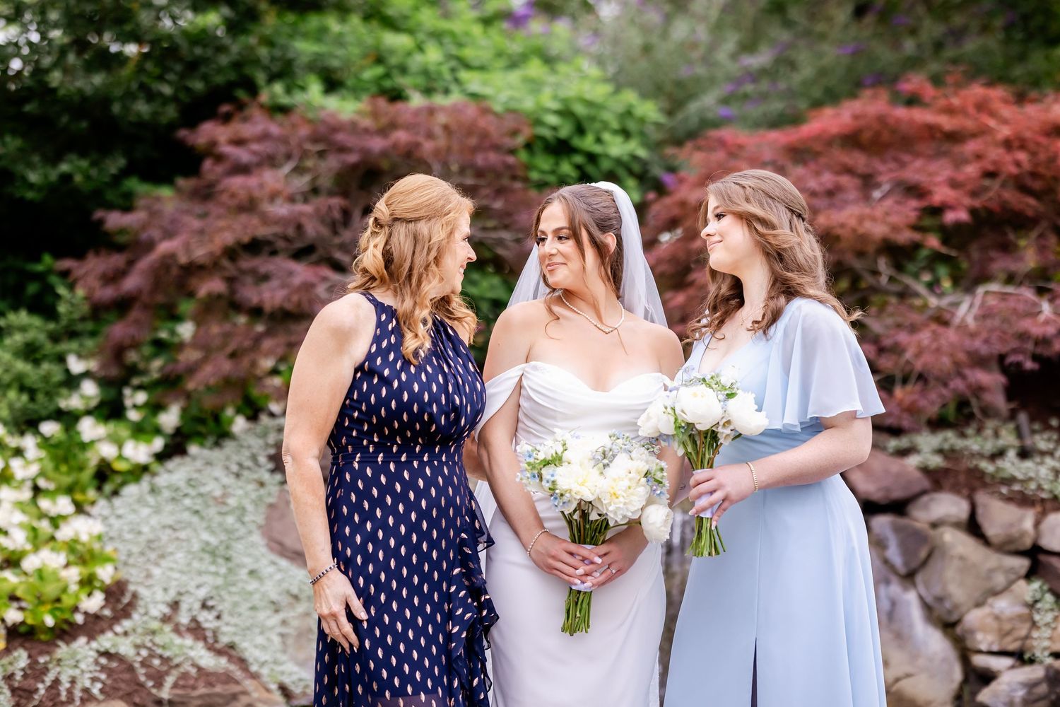 Three women in formal attire gather outdoors with bouquets against a backdrop of colorful Japanese maple trees.