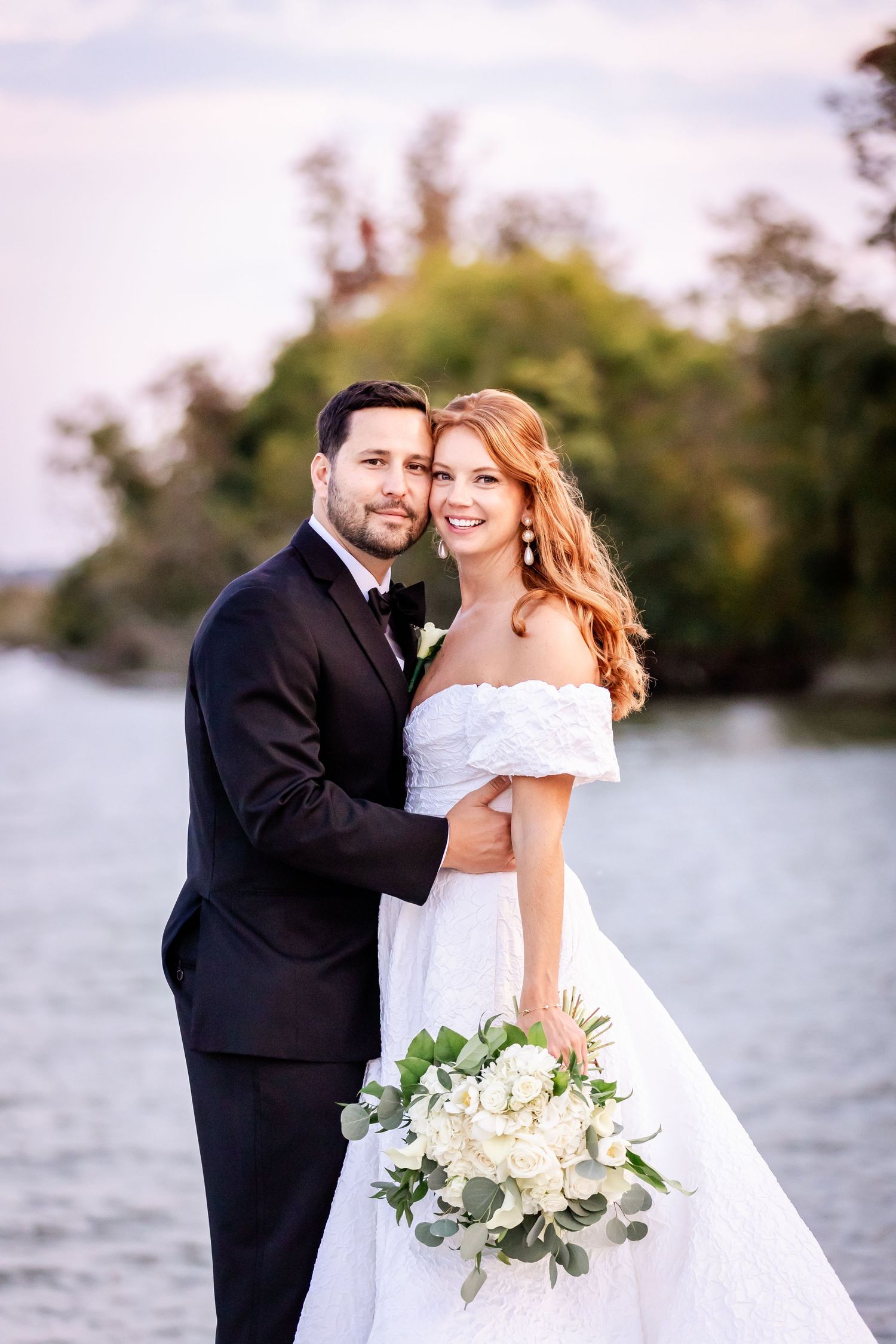 Newlyweds pose by the waterfront at sunset holding a white and green floral bouquet.