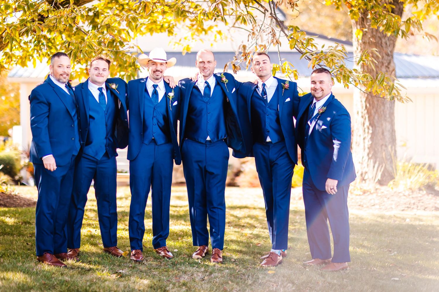 Members of a wedding party wearing coordinated navy blue suits and brown shoes pose together outdoors in autumn lighting.