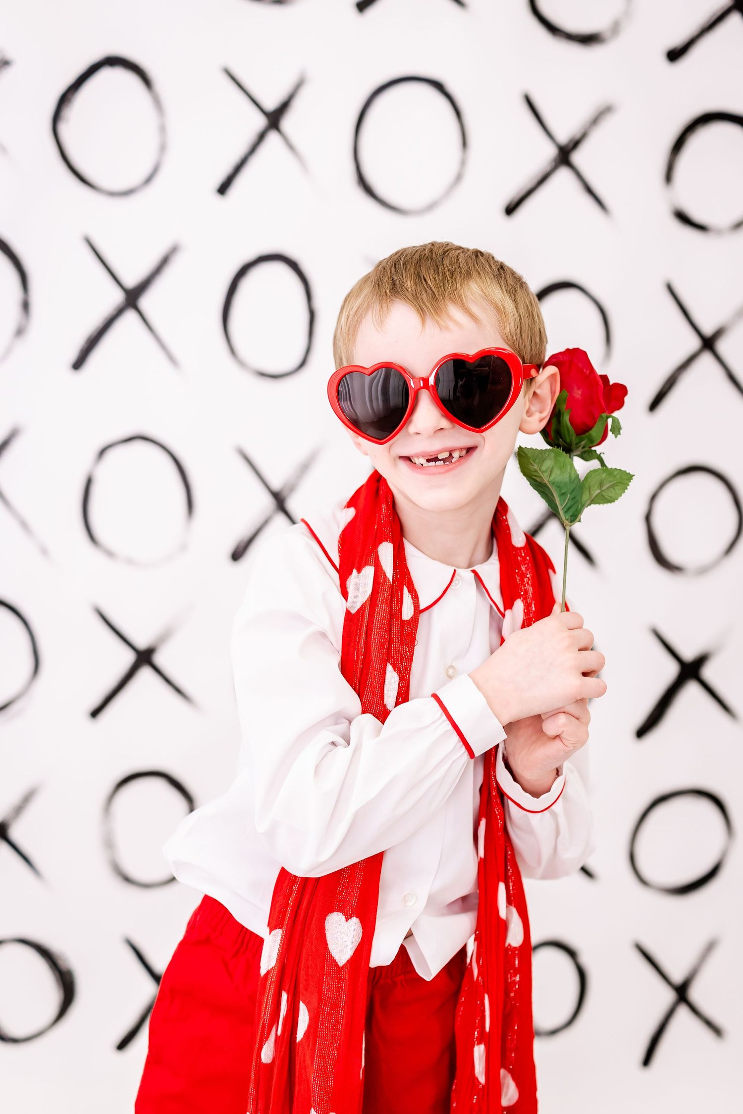 Whimsical Valentine's Day themed photo with heart sunglasses and red rose against XO pattern background.