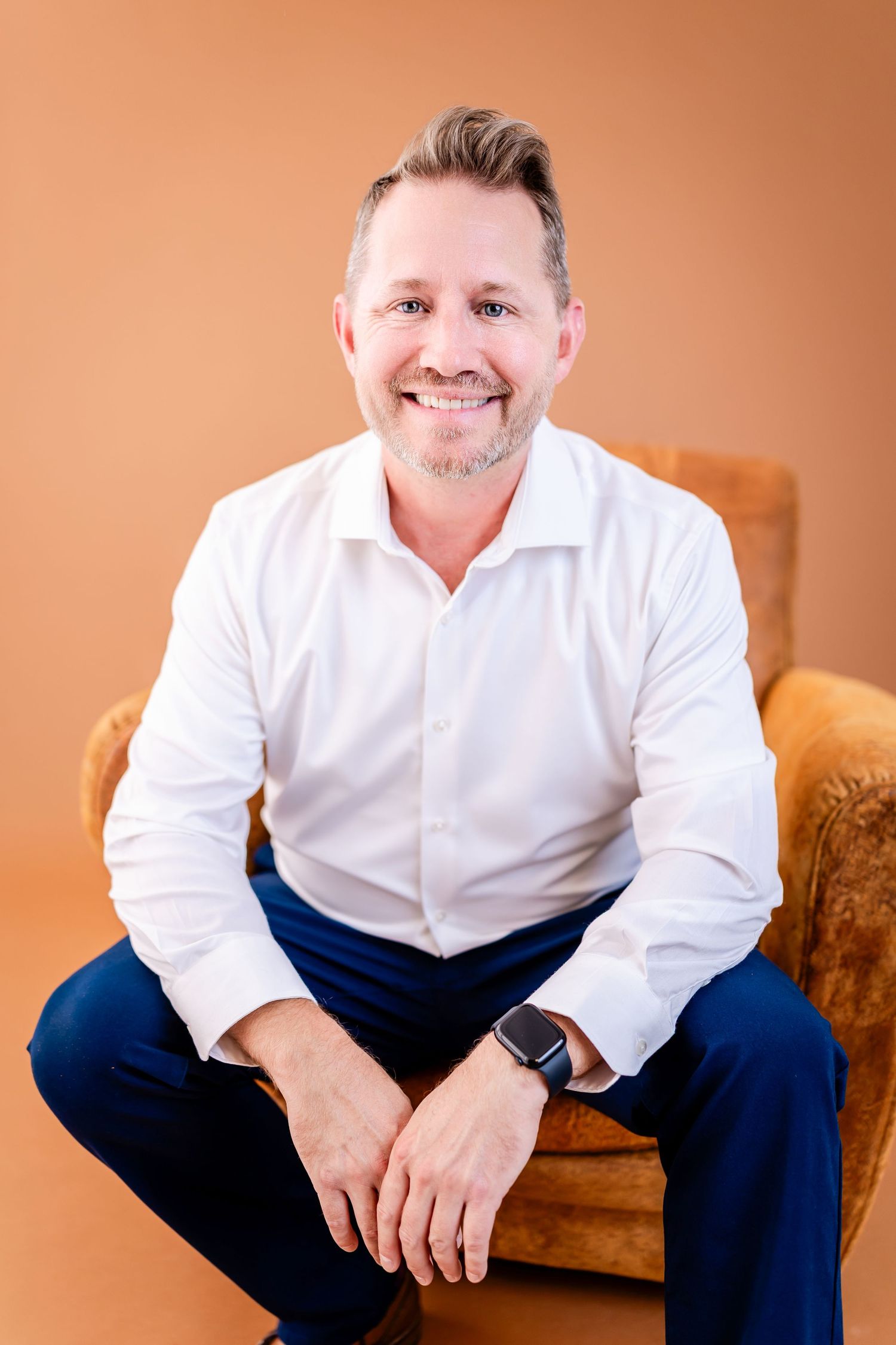 Smiling middle-aged man in white shirt and blue pants sitting in armchair against orange background.