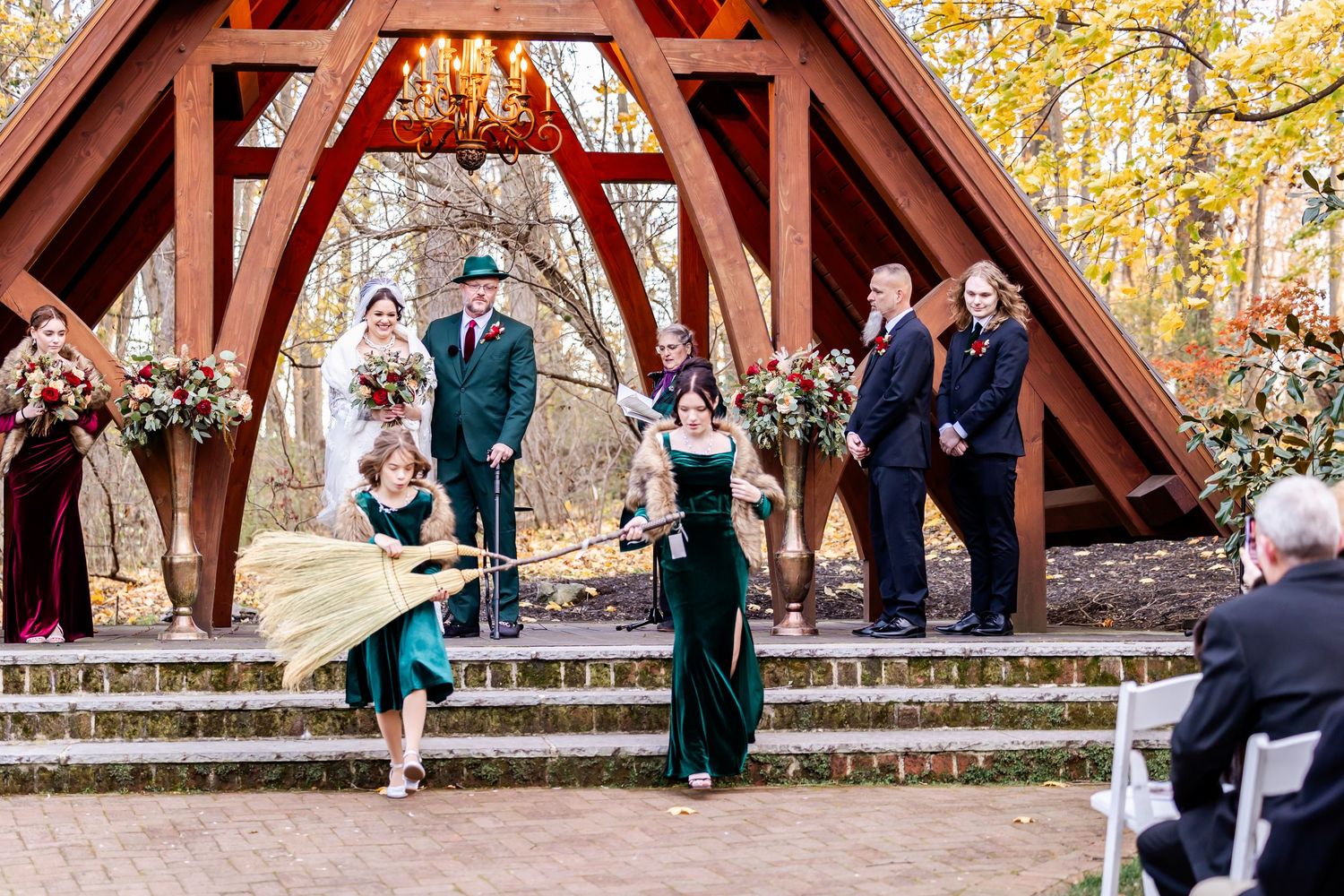 Wedding ceremony taking place under rustic wooden pavilion with guests seated on white chairs.