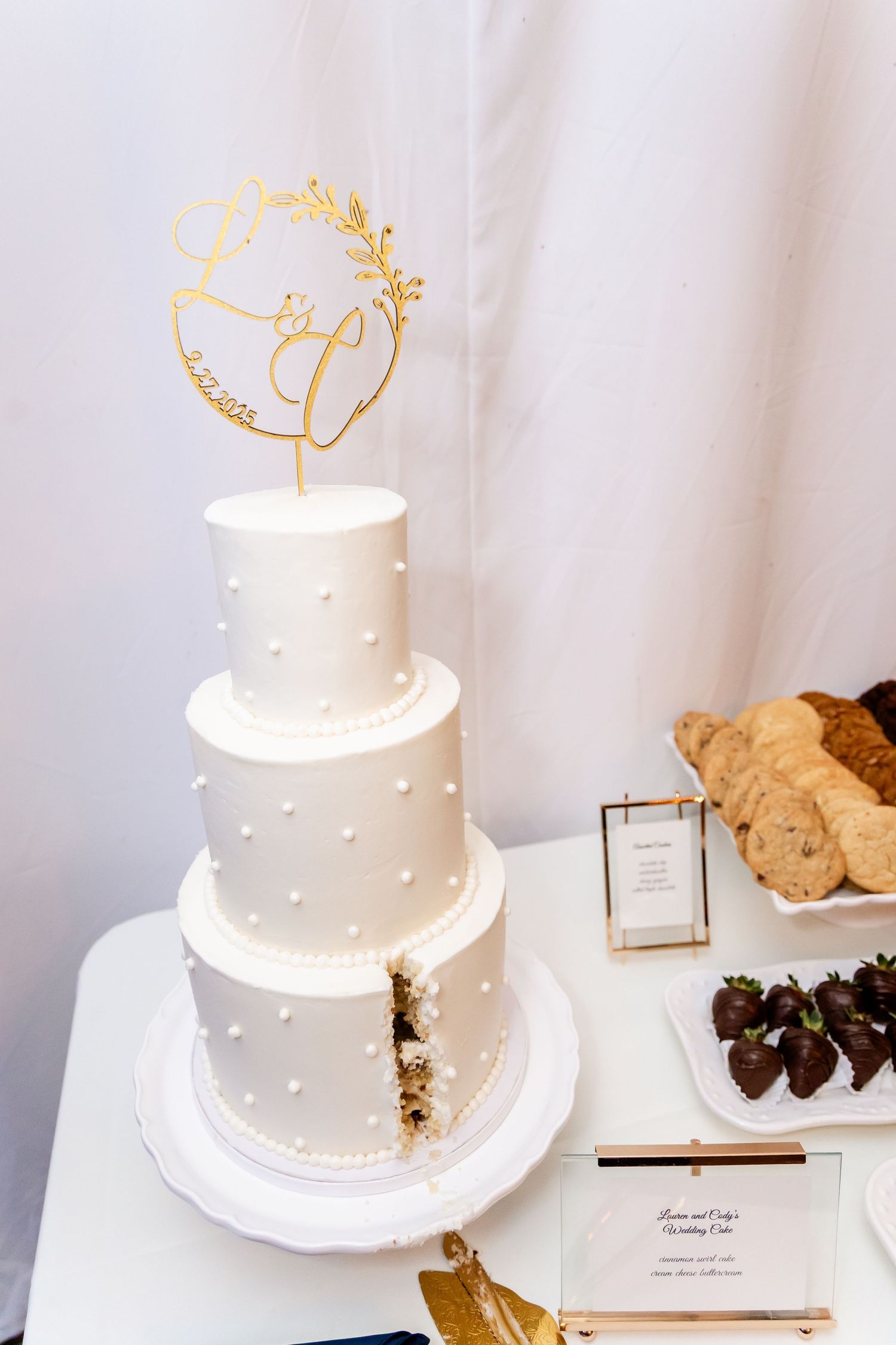 Elegant white tiered wedding cake with gold monogram topper and delicate pearl details.
