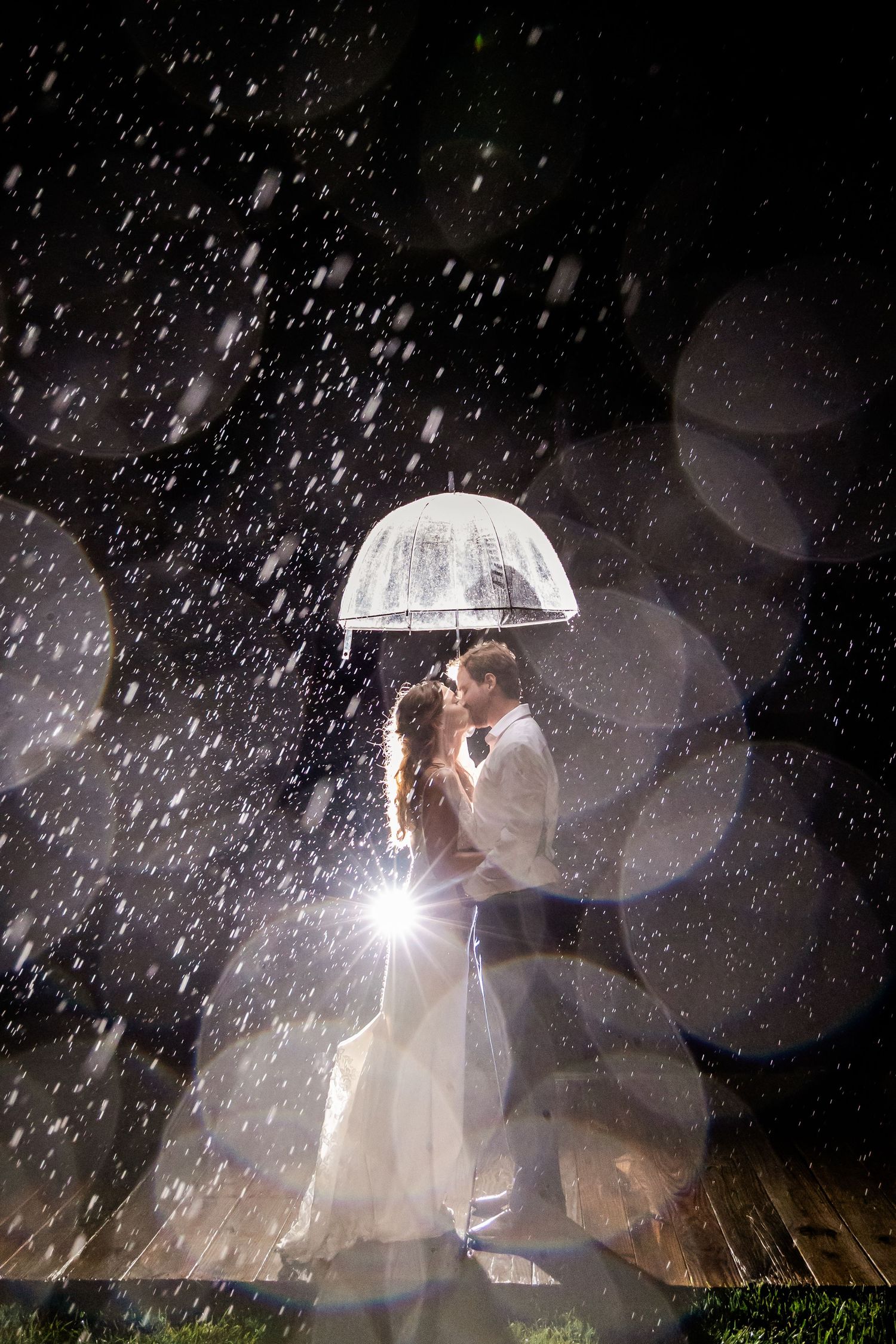 A romantic nighttime wedding photo showing silhouettes in the rain with an umbrella and dramatic lighting effects.