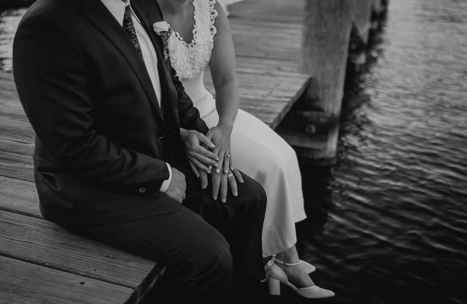Black and white photo of a wedding couple sitting on a dock, holding hands, wearing formal attire near water.