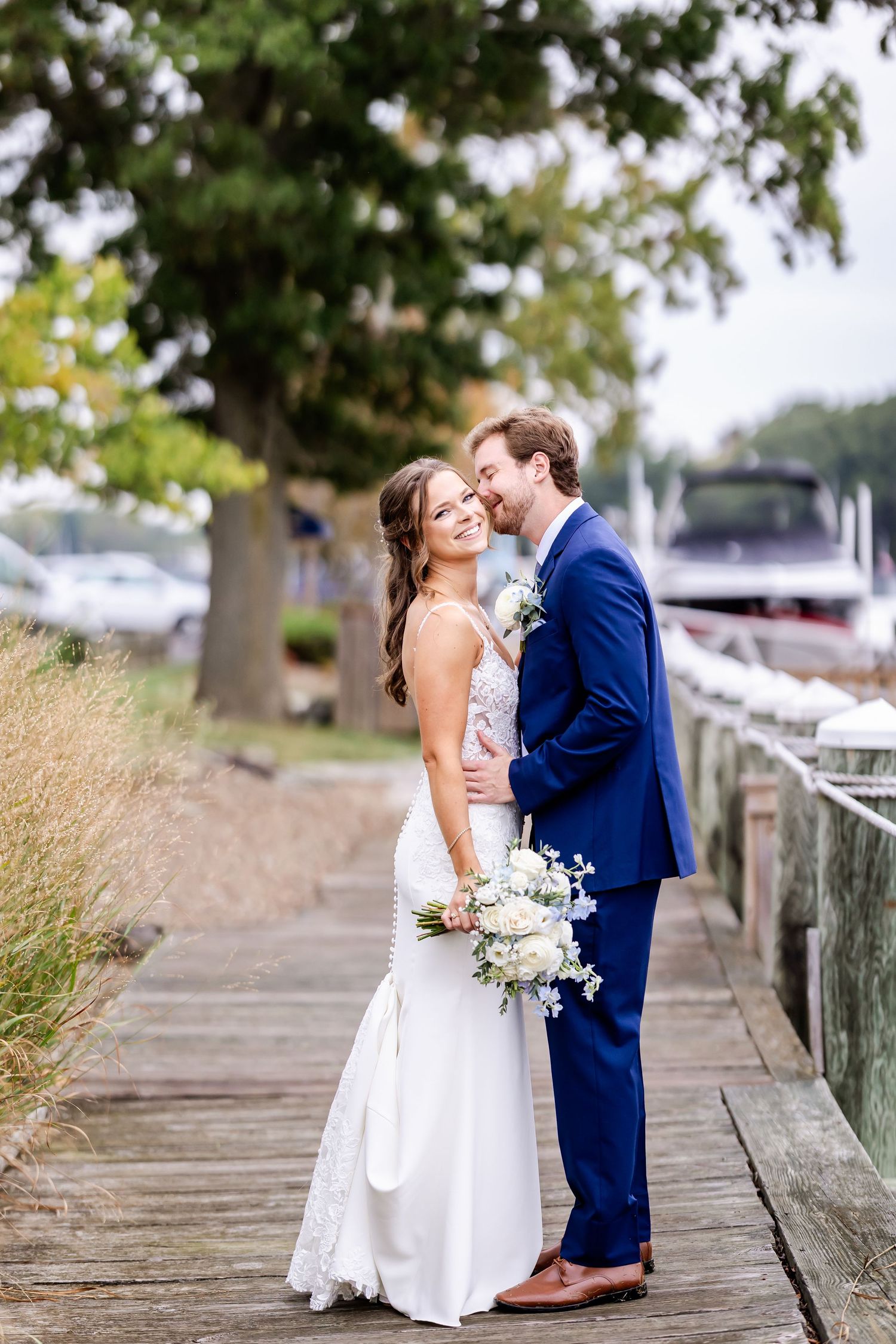 A romantic wedding couple poses on a wooden boardwalk by a marina with boats and trees in the background.