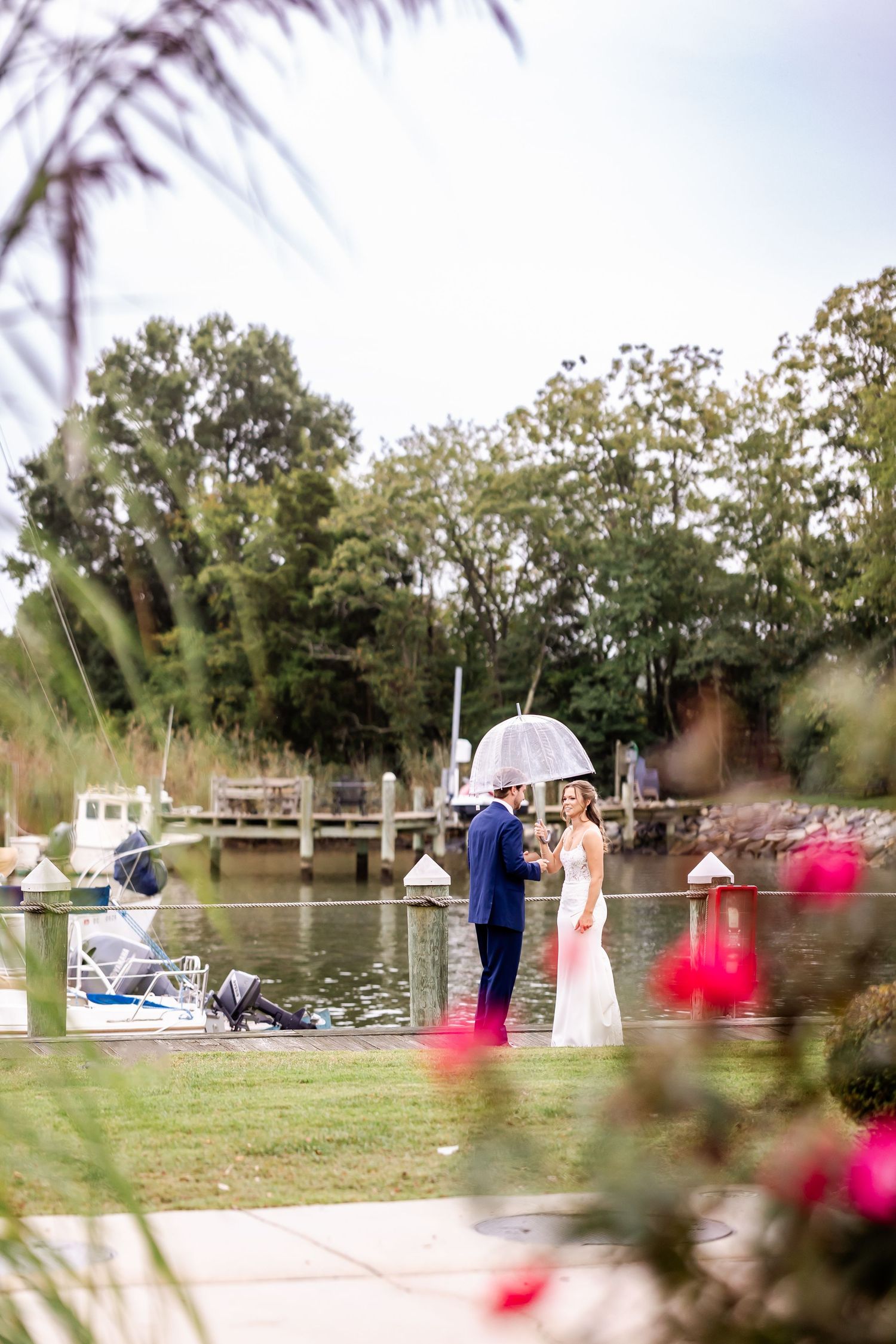 A romantic wedding moment by the waterfront dock with an umbrella and pink flowers in the foreground.