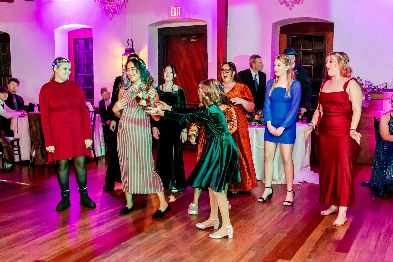 Wedding guests in colorful dresses dancing together on a pink-lit dance floor at an indoor reception.