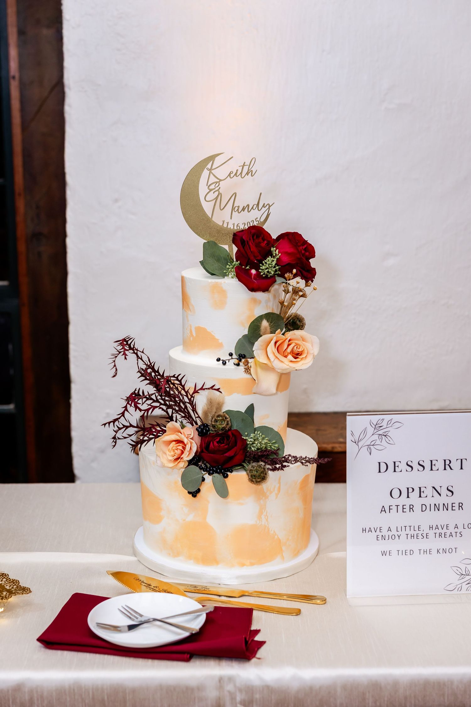 Elegant wedding dessert table with white cake decorated with red and peach roses and gold moon topper.