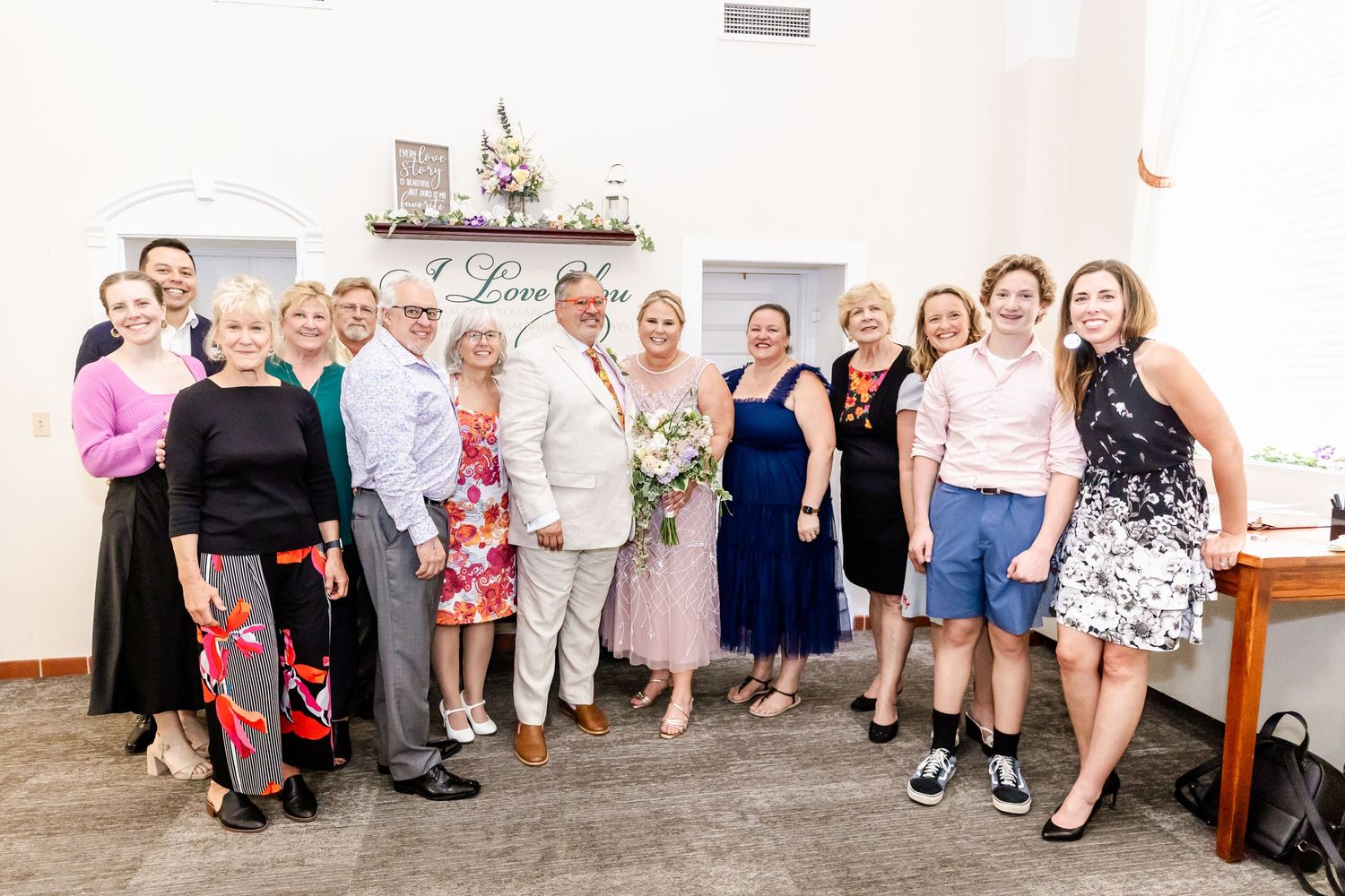 Group photo of wedding guests in formal attire gathered against a white wall.