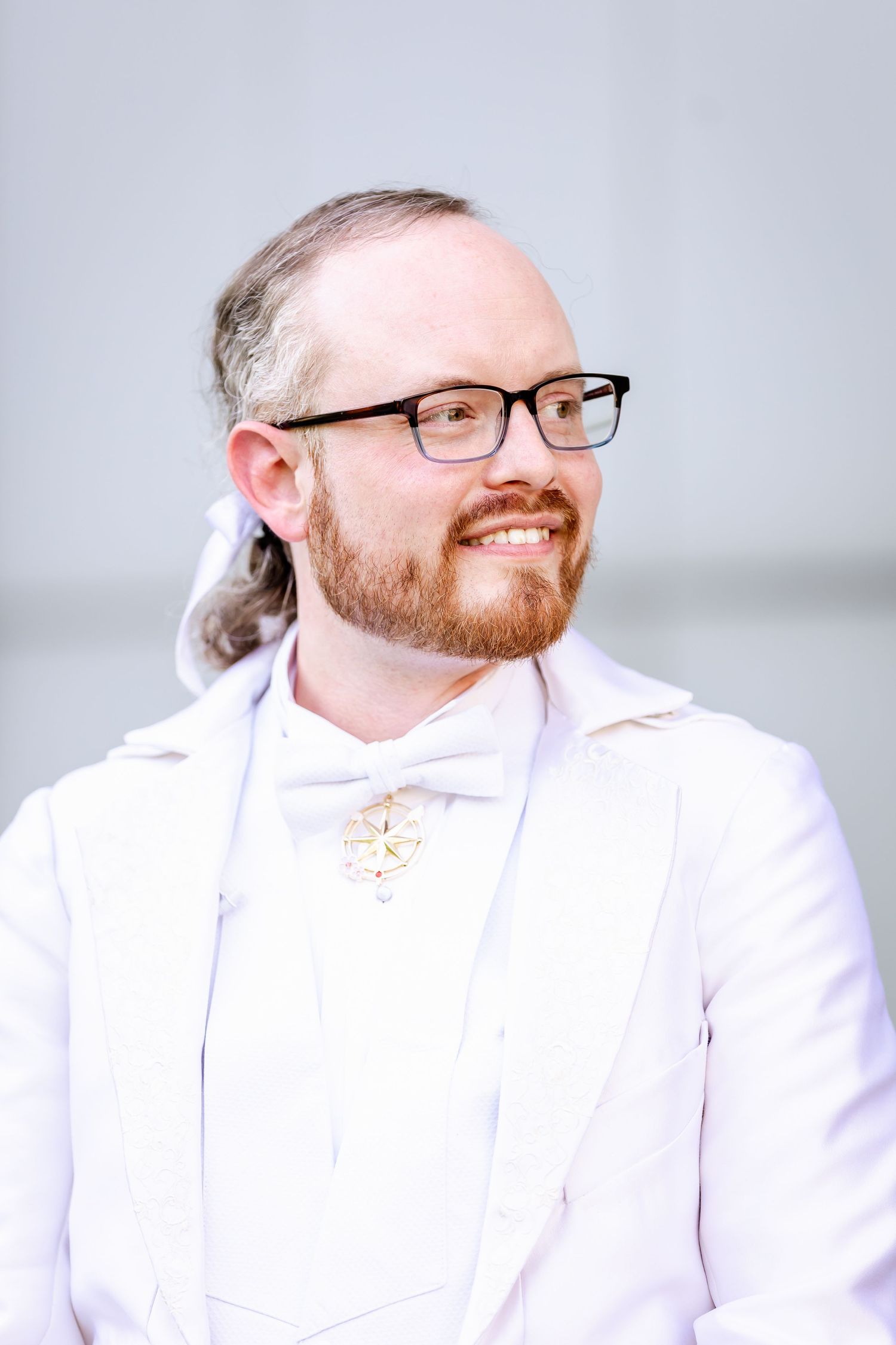 Person in white formal attire and glasses smiles warmly against a grey background.
