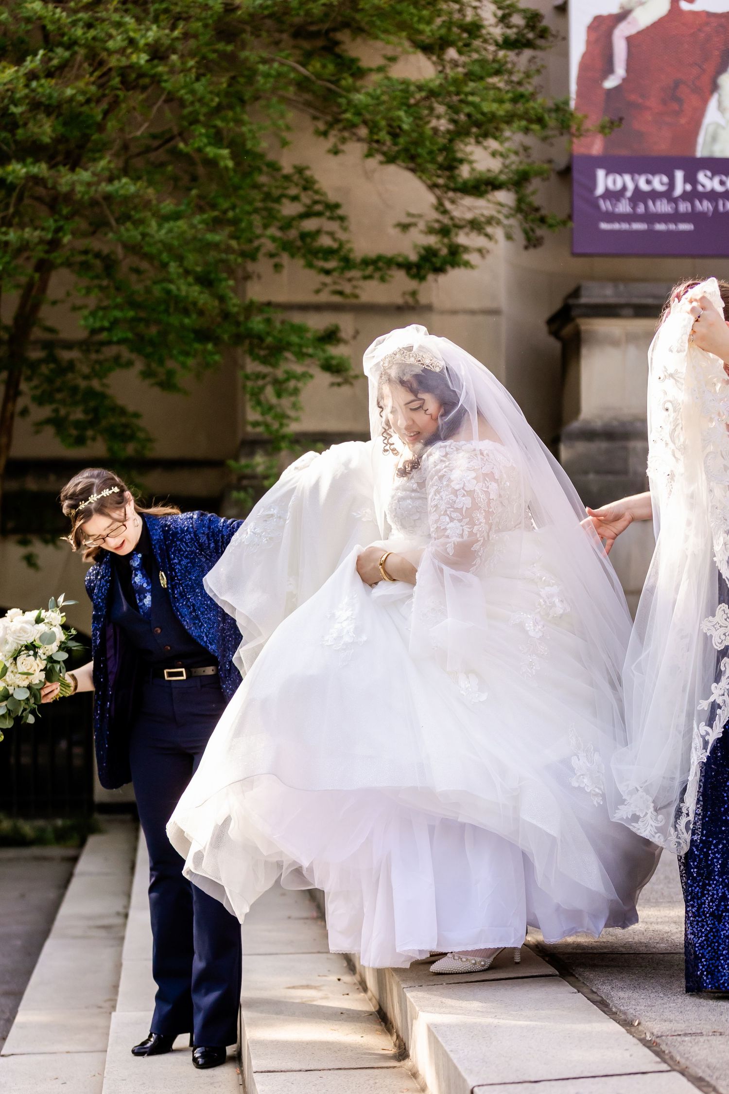 Wedding party members help bride navigate stairs in her flowing white gown while wearing navy blue attire at outdoor venue.