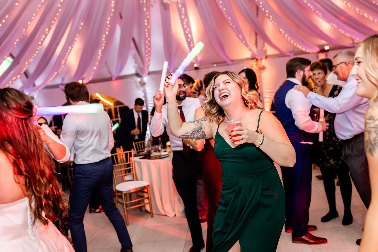 Energetic party guests dancing under pink-lit draped fabric ceiling at a wedding reception.