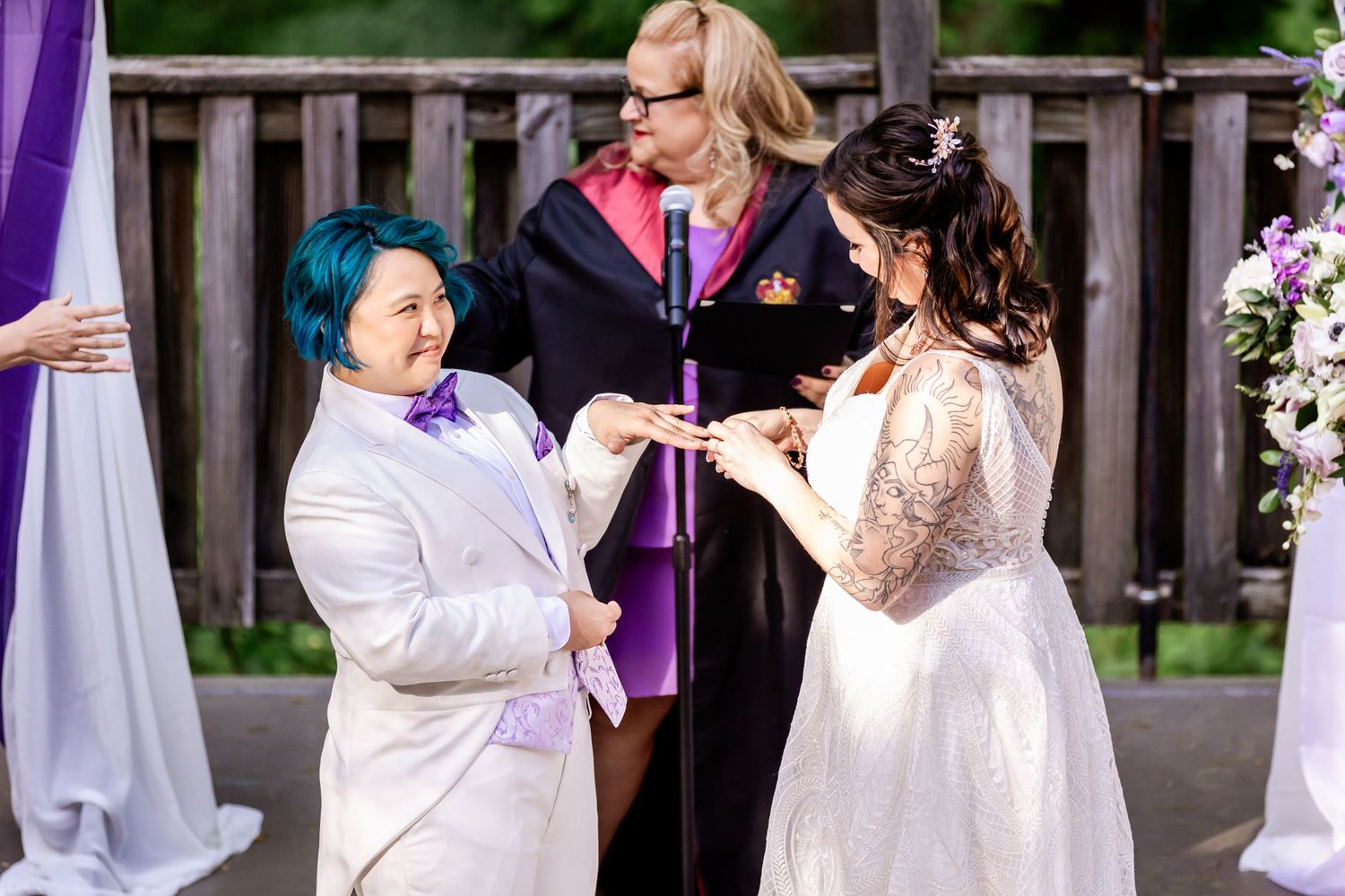 Two people exchange rings during their wedding ceremony with purple accents and flowers.