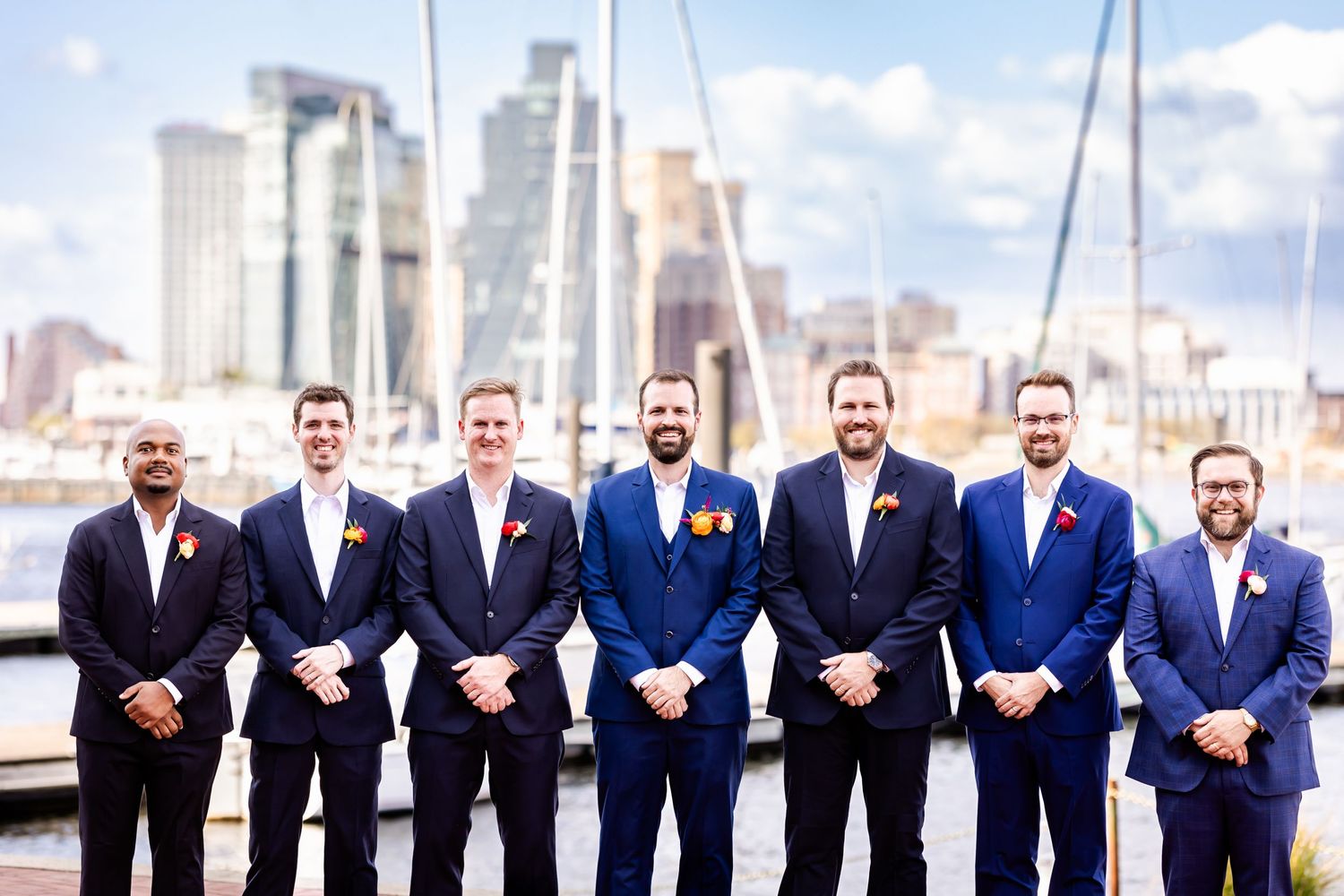 Group of men in formal suits stands together by a marina with city skyline in background.