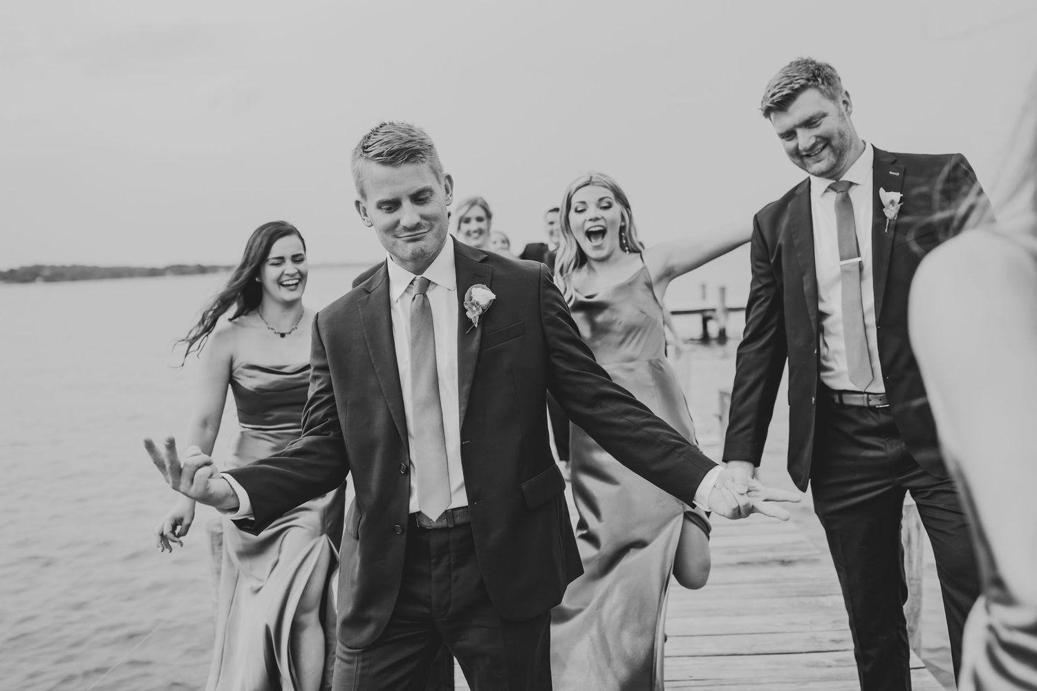 Black and white photo of a wedding party laughing together while walking on a dock by the water.