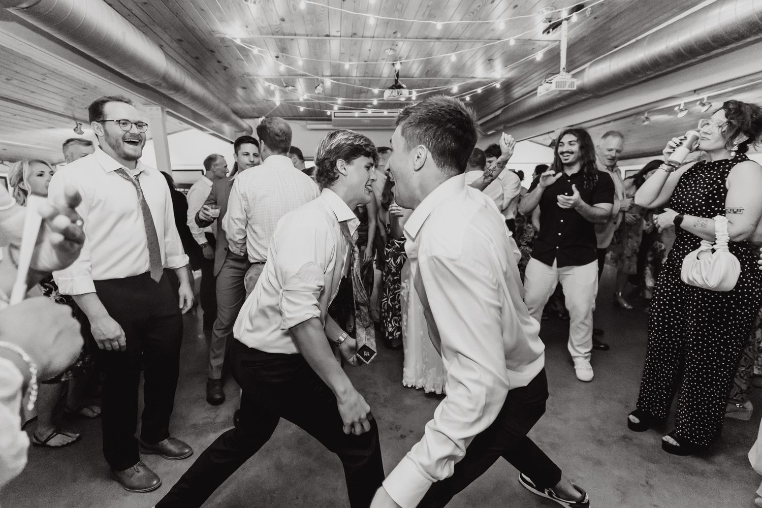 Black and white photo of guests dancing and celebrating at a lively wedding reception on a dance floor.