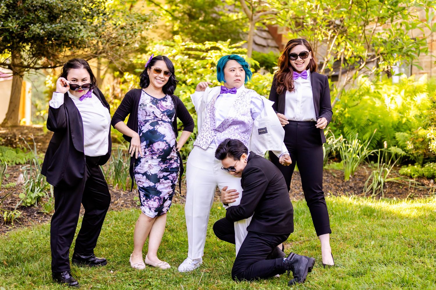 Playful wedding party posing dramatically in formal wear against green garden backdrop.