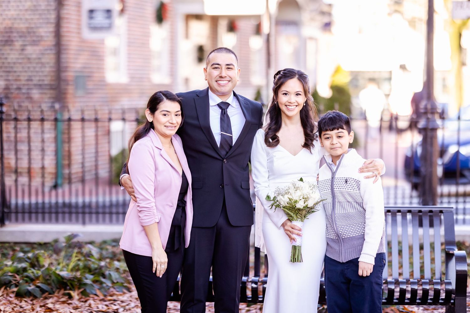 A bride in white dress poses with family members on a brick sidewalk during an autumn wedding.
