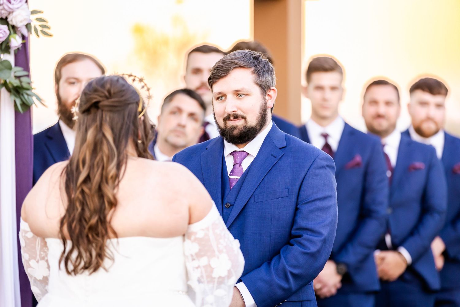 A wedding ceremony under an archway with bridesmaids in purple and groomsmen in navy suits.