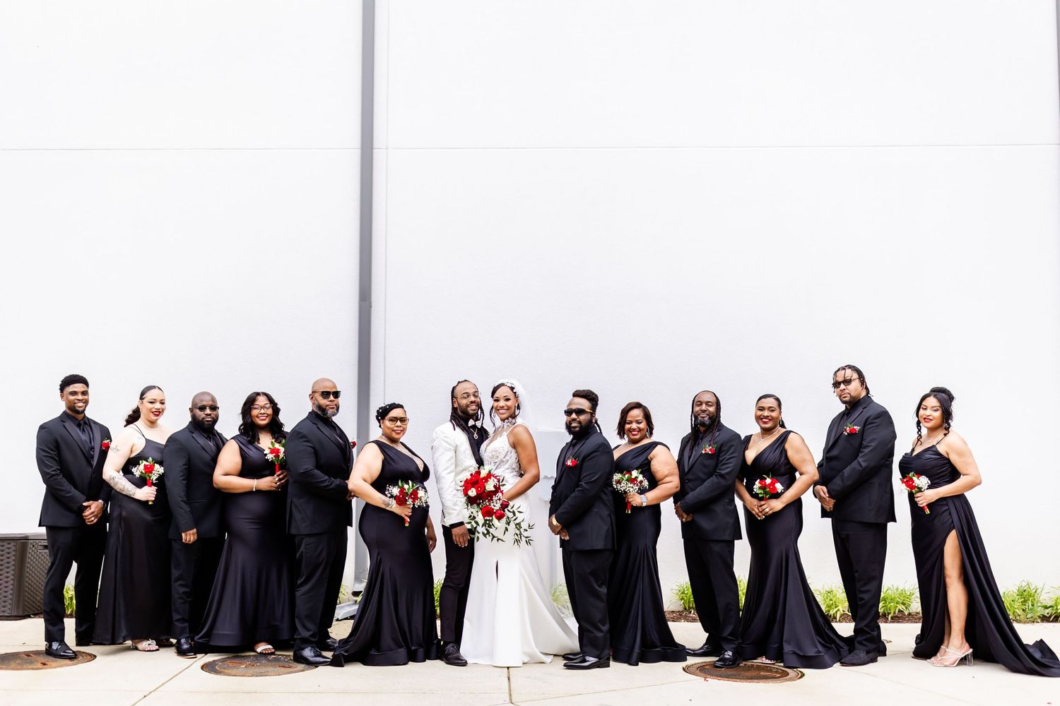 Full wedding party in formal attire poses against minimalist white wall.