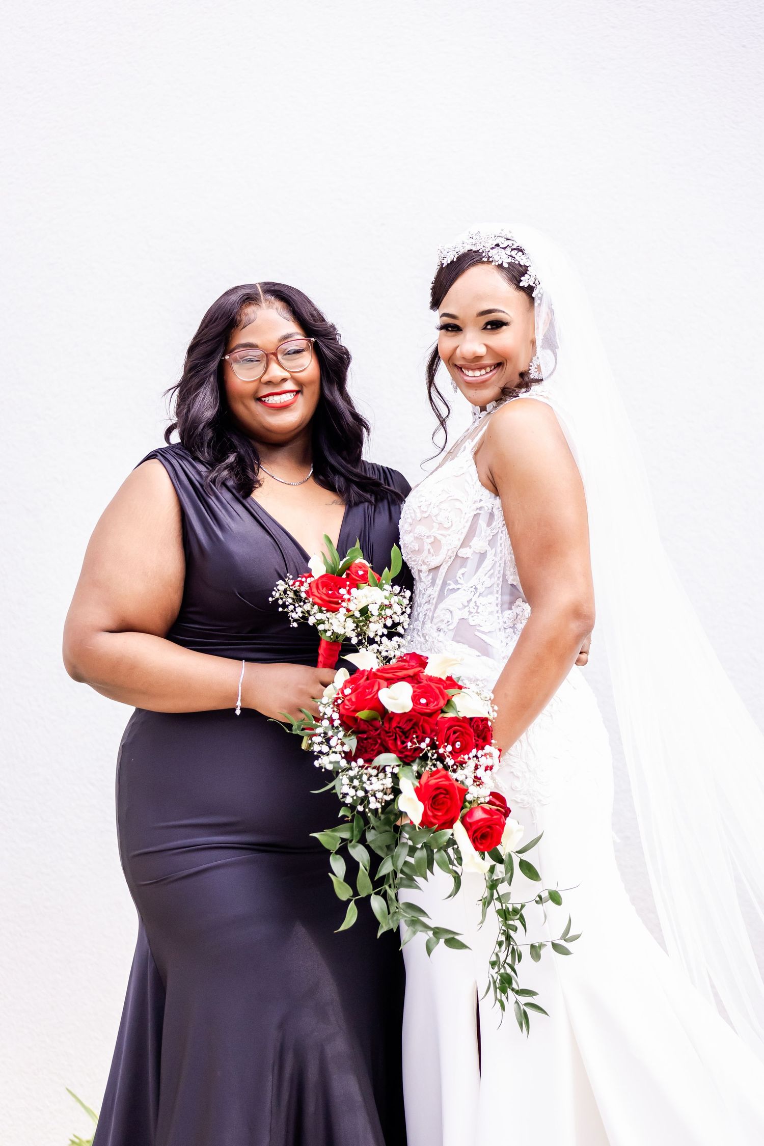 Two people pose with red rose bouquets, one in black and one in white wedding attire.