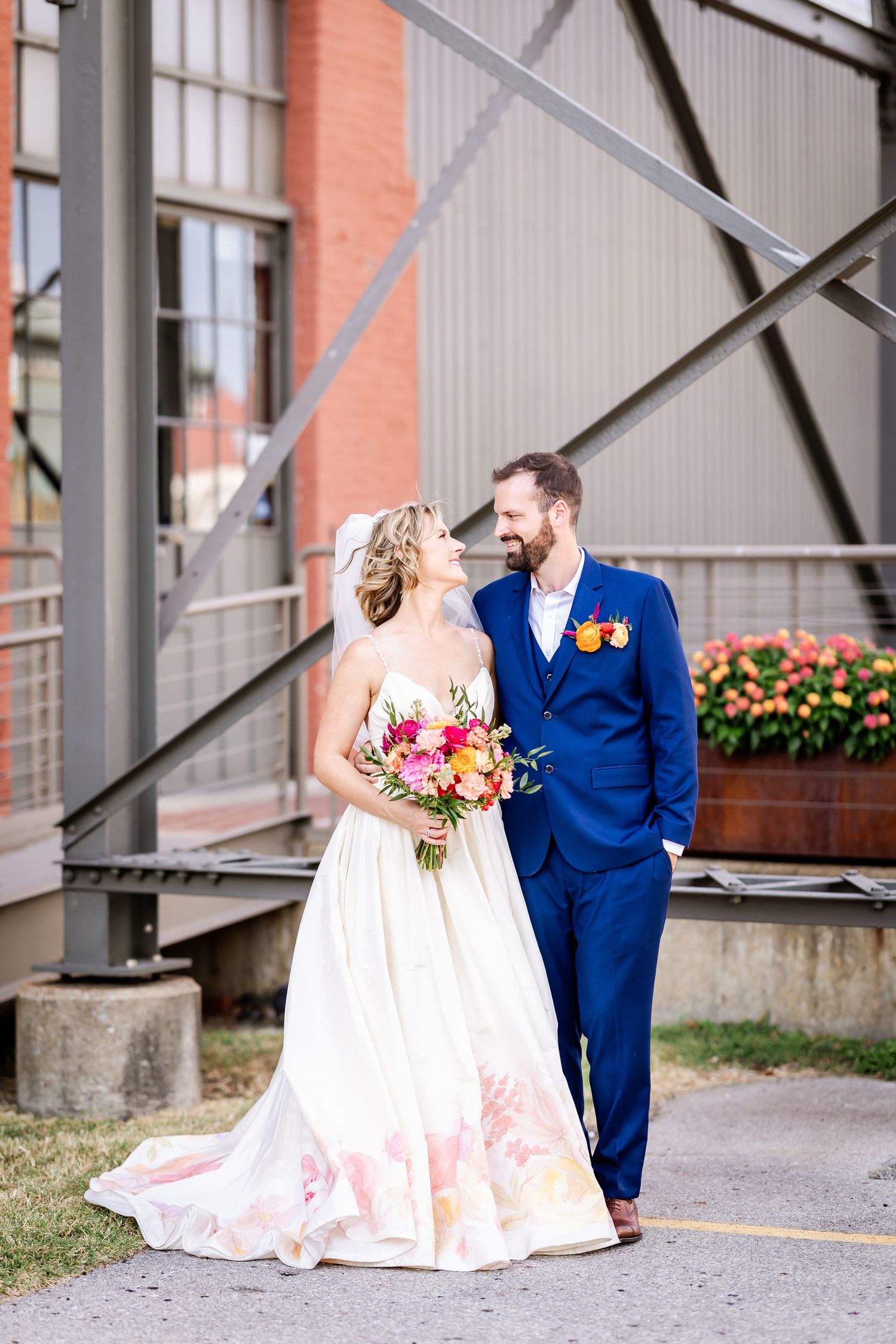 Elegant couple in wedding attire standing by industrial architecture with pink bouquet.