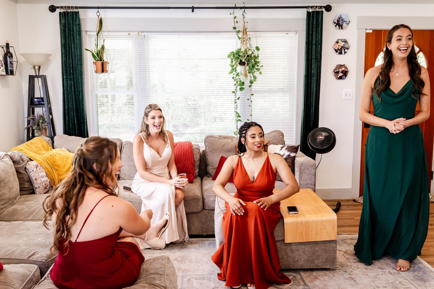 Wedding party members sit on couches in coordinated dresses of burgundy, green, and neutral colors.
