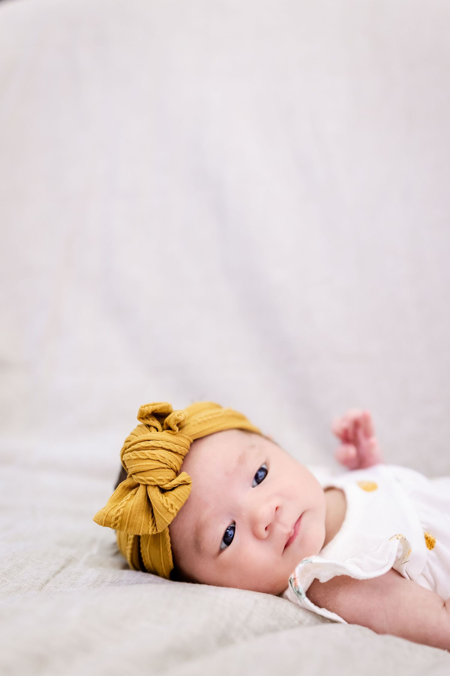 A baby with yellow headbow rests peacefully on white bedding during newborn photography session.
