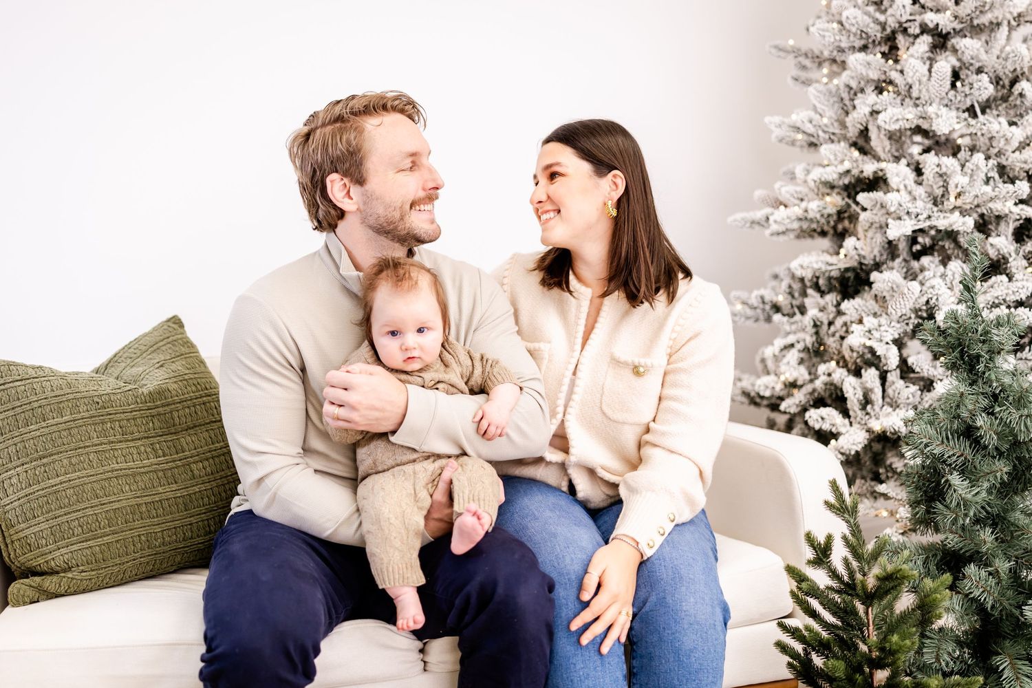 A young family sits together on a white couch near a decorated Christmas tree during a holiday photoshoot.