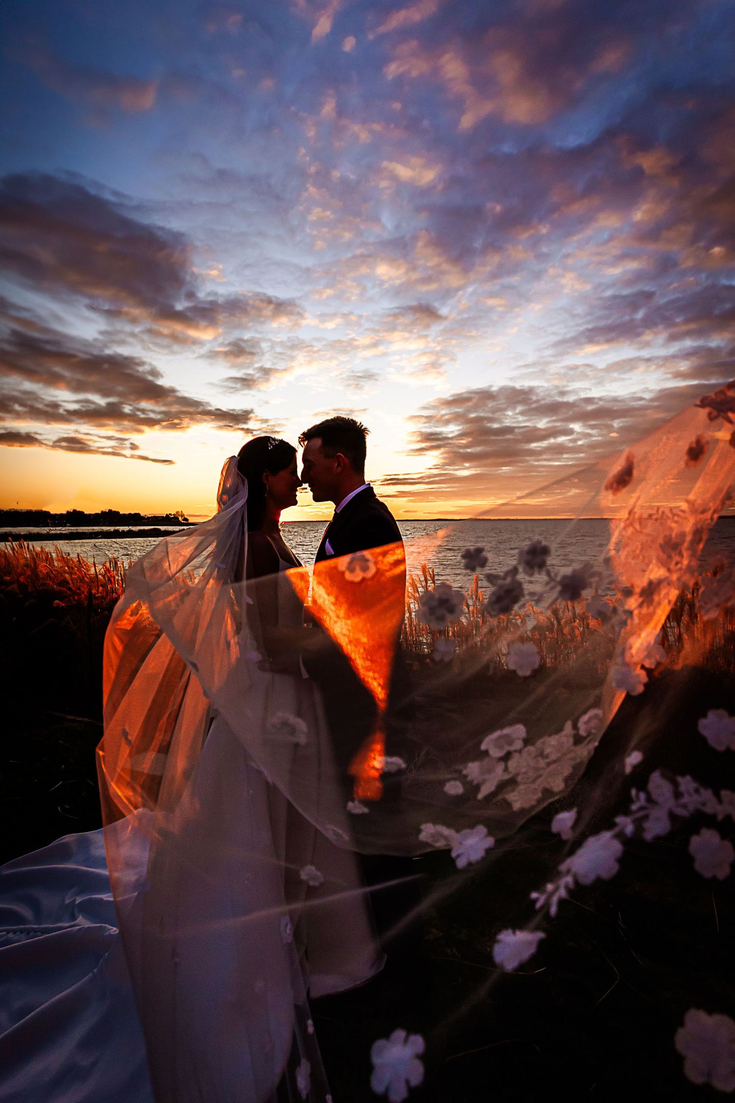 A romantic sunset silhouette of a couple with a flowing veil creates a dramatic wedding photo series against vibrant orange skies.