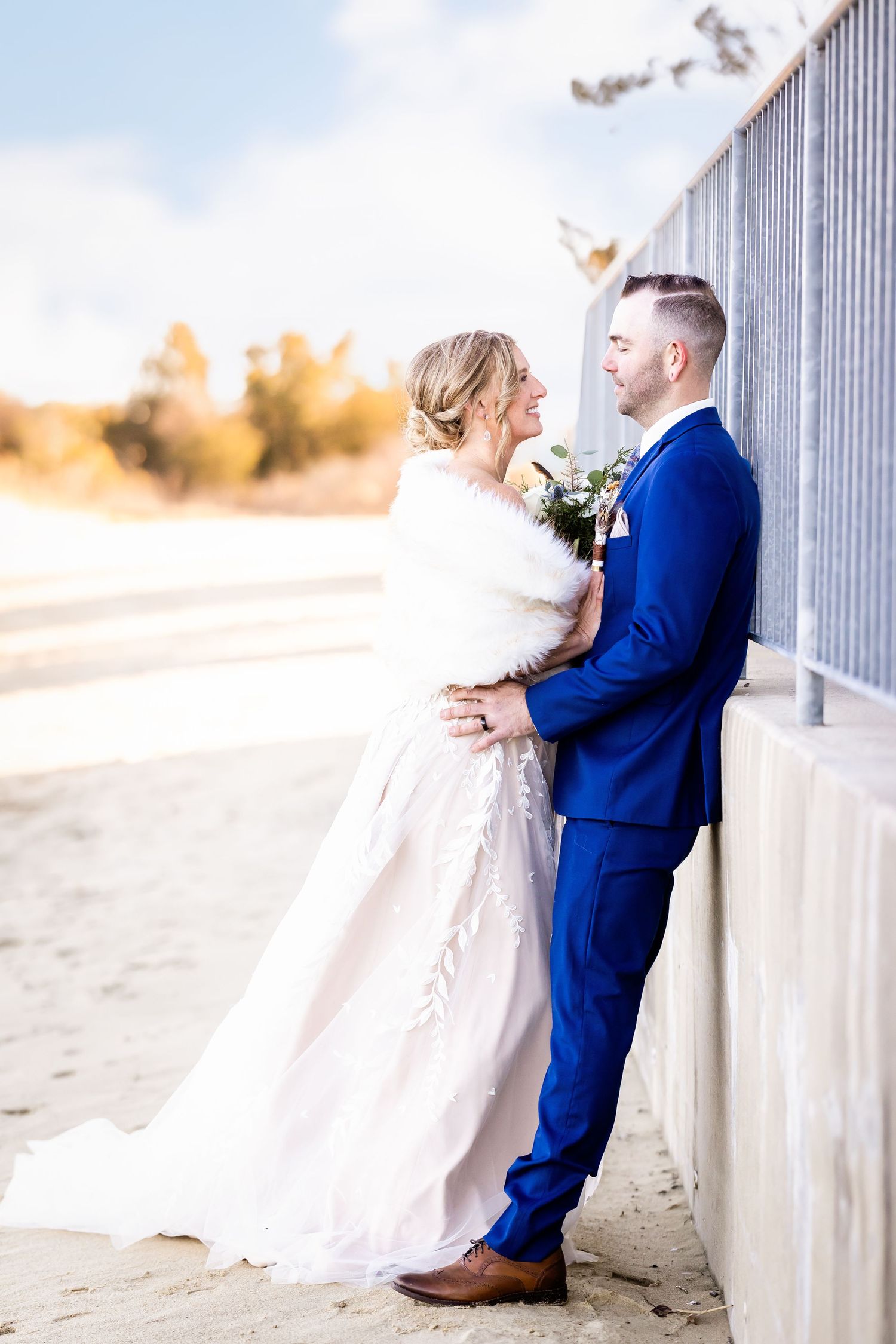 A couple in wedding attire poses together against a wall, with the bride wearing white and groom in a royal blue suit.