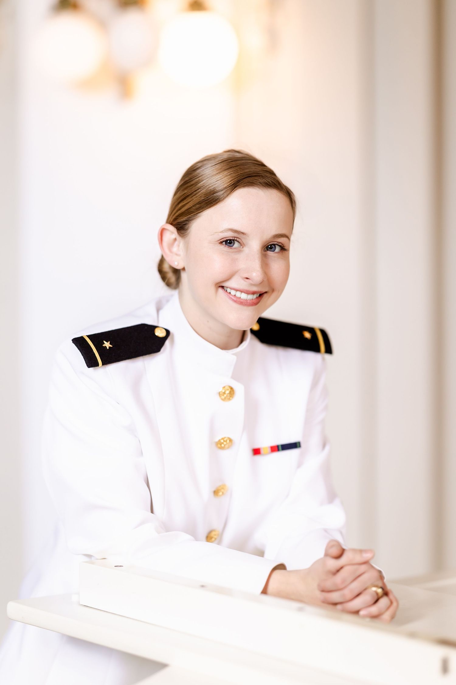 Naval officer in dress white uniform with rank insignia and medals sits at a desk with a bright smile.