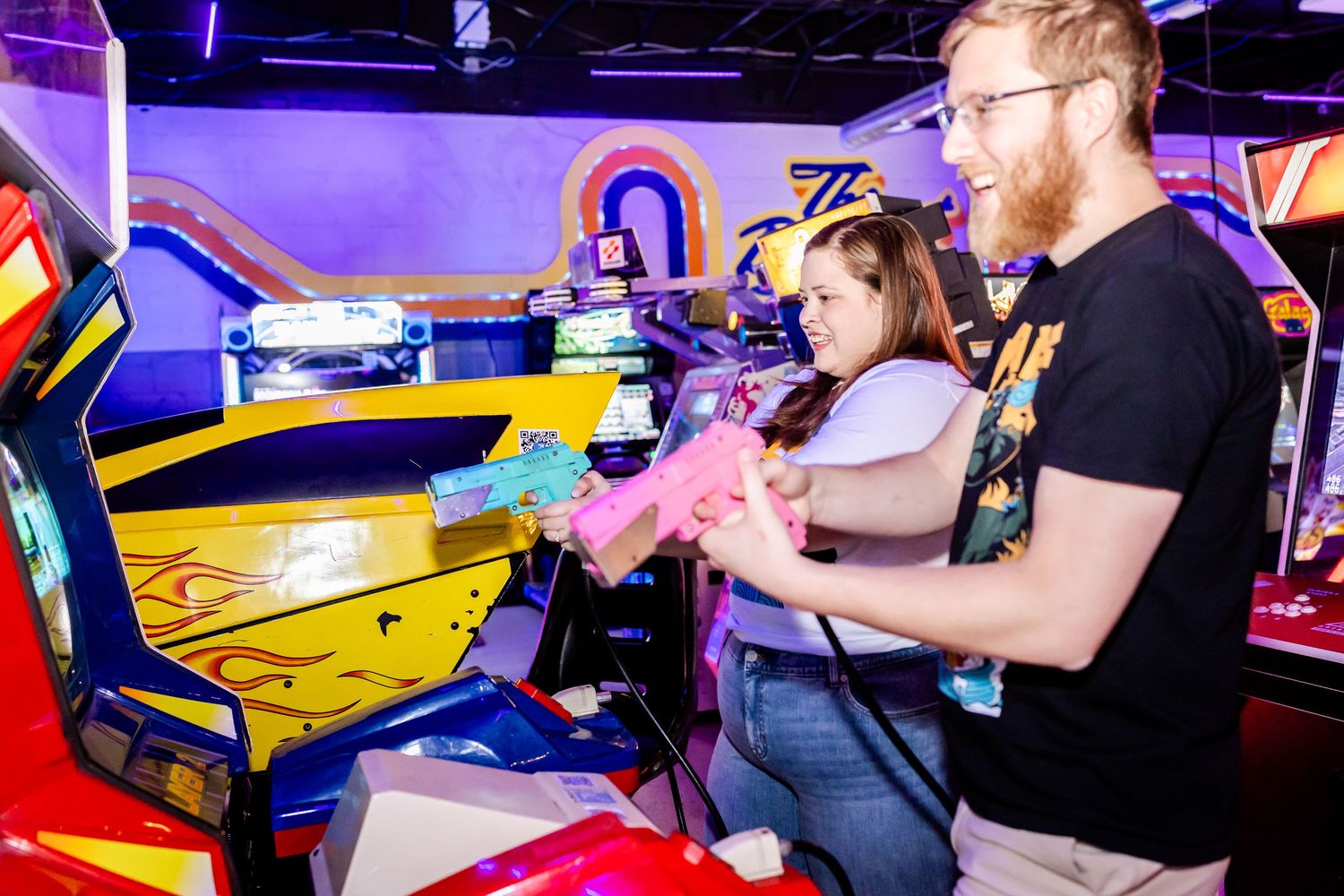 Two people playing a colorful arcade racing game together in a vibrant neon-lit gaming venue.