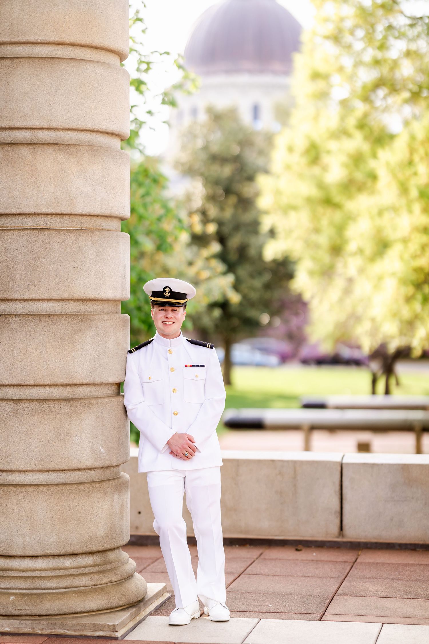 Naval officer in dress whites standing beside classical column with dome visible in background.