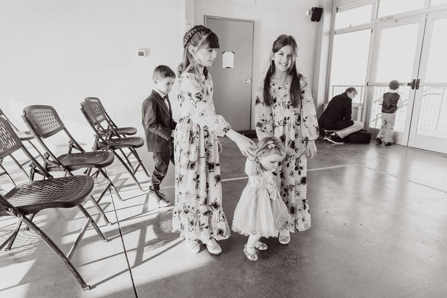 Wedding party members in floral dresses gather in a room with folding chairs.