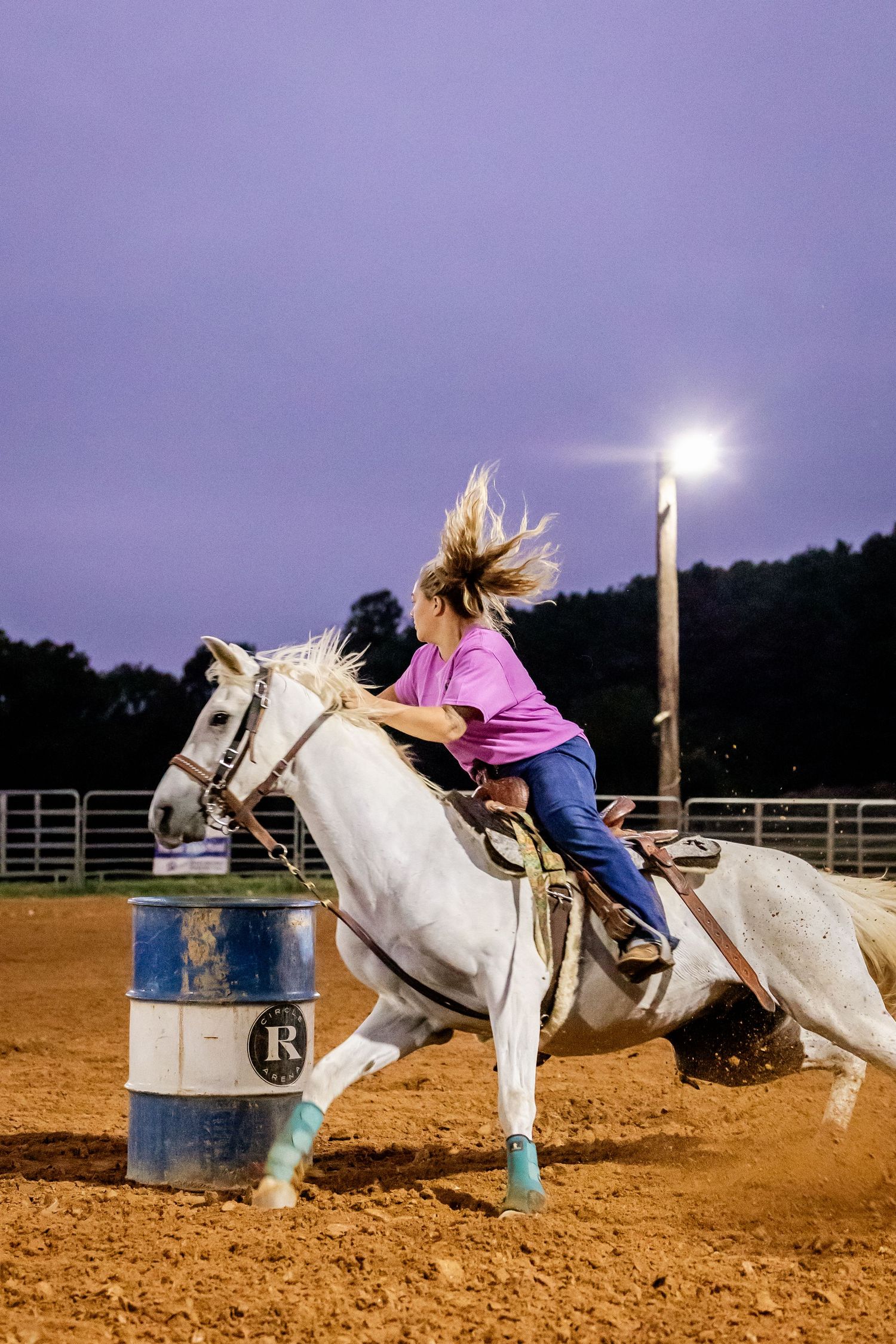 White horse and rider compete in barrel racing under arena lights at dusk.