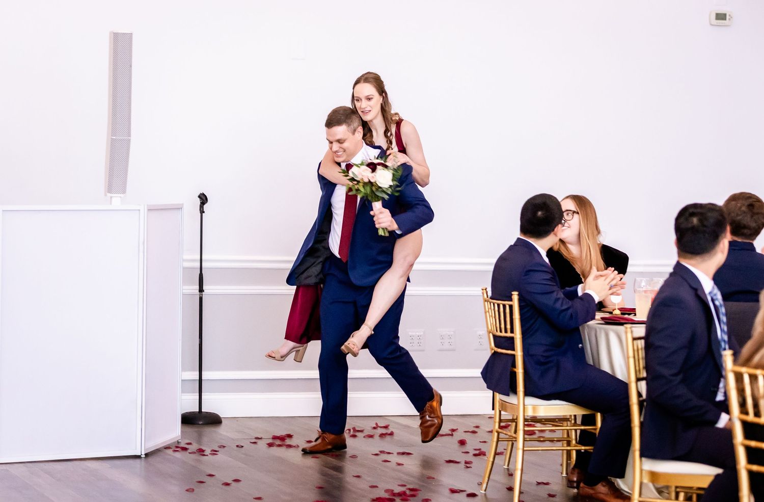 Wedding celebration in a bright indoor venue with guests seated on gold chairs.