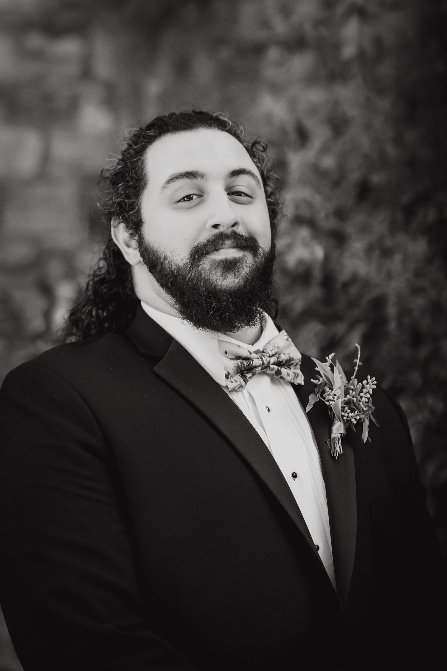 A formal portrait in black and white of someone wearing a tuxedo with a boutonniere and bowtie.